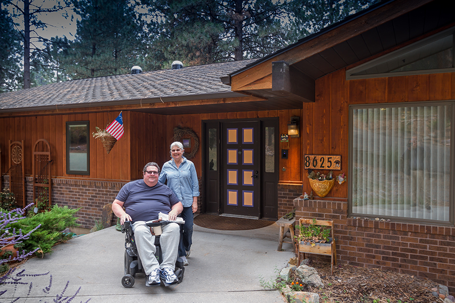 A smiling couple wearing blue shirts and tinted glasses pose on the accessible pathway in front of their house. The woman places her hand on the man's back, while he sits in a wheelchair holding a newspaper in his left hand and a cup of coffee between his legs.